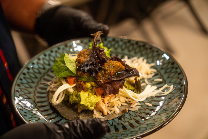Vibrant Mexican-inspired open taco on a blue patterned ceramic plate, with a tortilla topped with guacamole, diced tomato, shredded cheese, rice and mixed salad greens, presented by gloved hands.