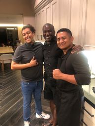 Three smiling friends posing in a modern home kitchen with white cabinets and dark wood floors — casual gathering, one wearing an apron and a dining table with guests in the background.