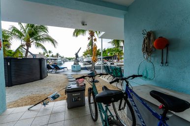 Covered tropical waterfront patio overlooking a canal with palm trees and docked boats; bicycles, fishing gear, cooler and lounge chairs in the foreground.