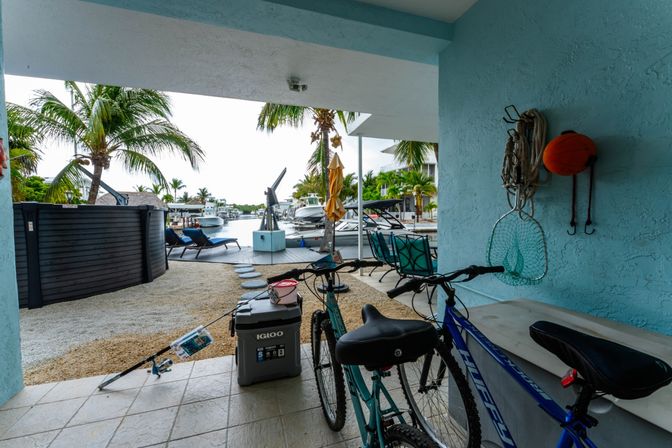 Covered tropical waterfront patio overlooking a canal with palm trees and docked boats; bicycles, fishing gear, cooler and lounge chairs in the foreground.