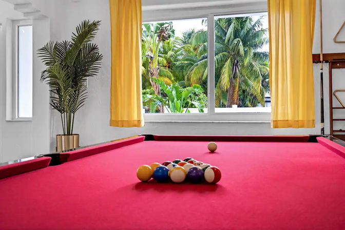 Red-felt pool table with racked billiard balls in a bright indoor game room; yellow curtains frame a large window overlooking lush tropical palm trees.
