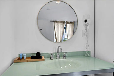 Minimal modern bathroom vanity with a round mirror reflecting sunlit curtains, mint glass countertop, chrome faucet, wooden tray with toiletries, and a wall-mounted hair dryer.