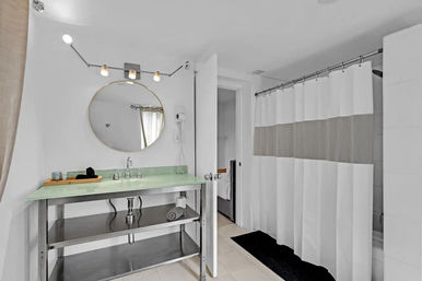 Bright modern bathroom interior with frosted glass countertop and sink, round gold-framed mirror, stainless-steel open-shelf vanity, three-bulb light fixture, white-and-gray shower curtain and black bath mat