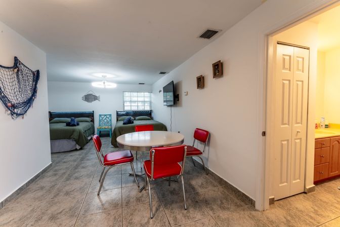 Beach-themed studio apartment with two green beds, wall-mounted TV, round table with retro red diner chairs, tiled floor and small bathroom off to the side