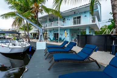 Tropical waterfront vacation home with a light-blue two-story house and balcony, docked white motorboat, palm trees and a row of blue cushioned lounge chairs on a wooden dock with an above-ground pool and outdoor dining area nearby.