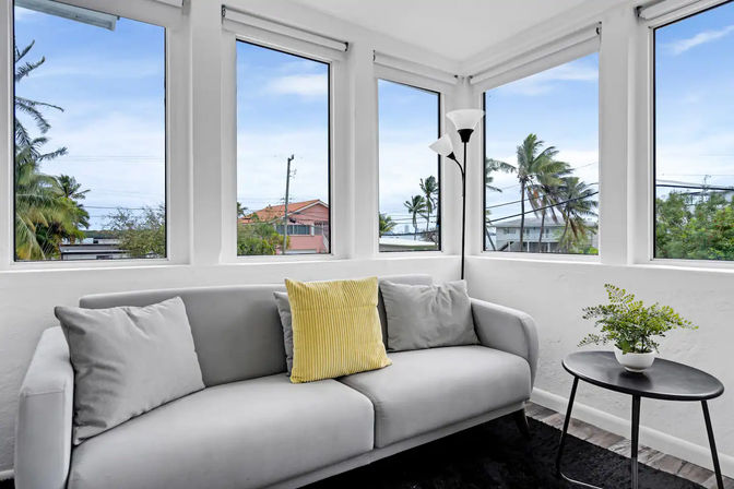 Sunlit coastal corner living room with large bay windows framing palm trees and rooftops, minimalist gray sofa with a yellow accent pillow, black floor lamp and round side table with potted plant.