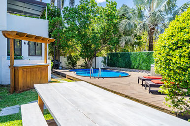 Sunny tropical backyard pool deck with round blue pool, wooden bar and picnic table in foreground, red-cushioned lounge chairs, palm trees and green privacy hedge.