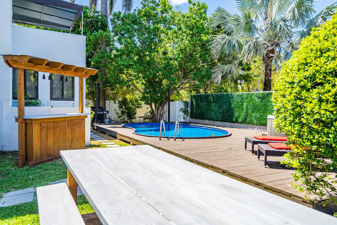 Sunny tropical backyard pool deck with round blue pool, wooden bar and picnic table in foreground, red-cushioned lounge chairs, palm trees and green privacy hedge.