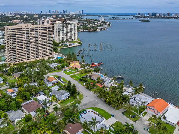 Aerial view of a coastal bayfront neighborhood with tall waterfront condos, palm-lined single-family homes, docks and a red crane boat in the bay, with a distant city skyline on the horizon.