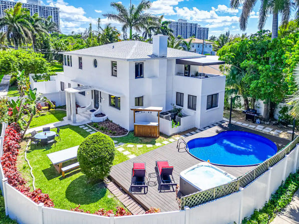 Sunny aerial view of a modern two-story white house with tropical landscaping, fenced backyard, deck with lounge chairs, kidney-shaped pool and hot tub.