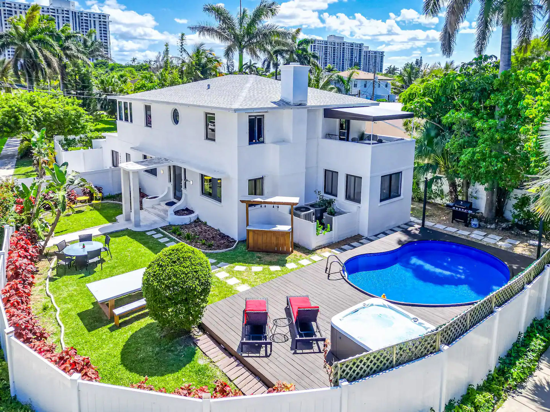 Sunny aerial view of a modern two-story white house with tropical landscaping, fenced backyard, deck with lounge chairs, kidney-shaped pool and hot tub.