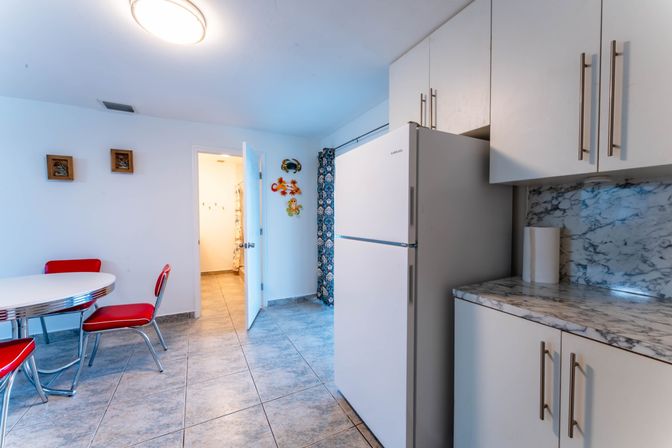 Bright kitchen dining nook with retro red diner chairs around a round white table, white refrigerator and cabinets, marble-look countertop and backsplash, gray tile floor.