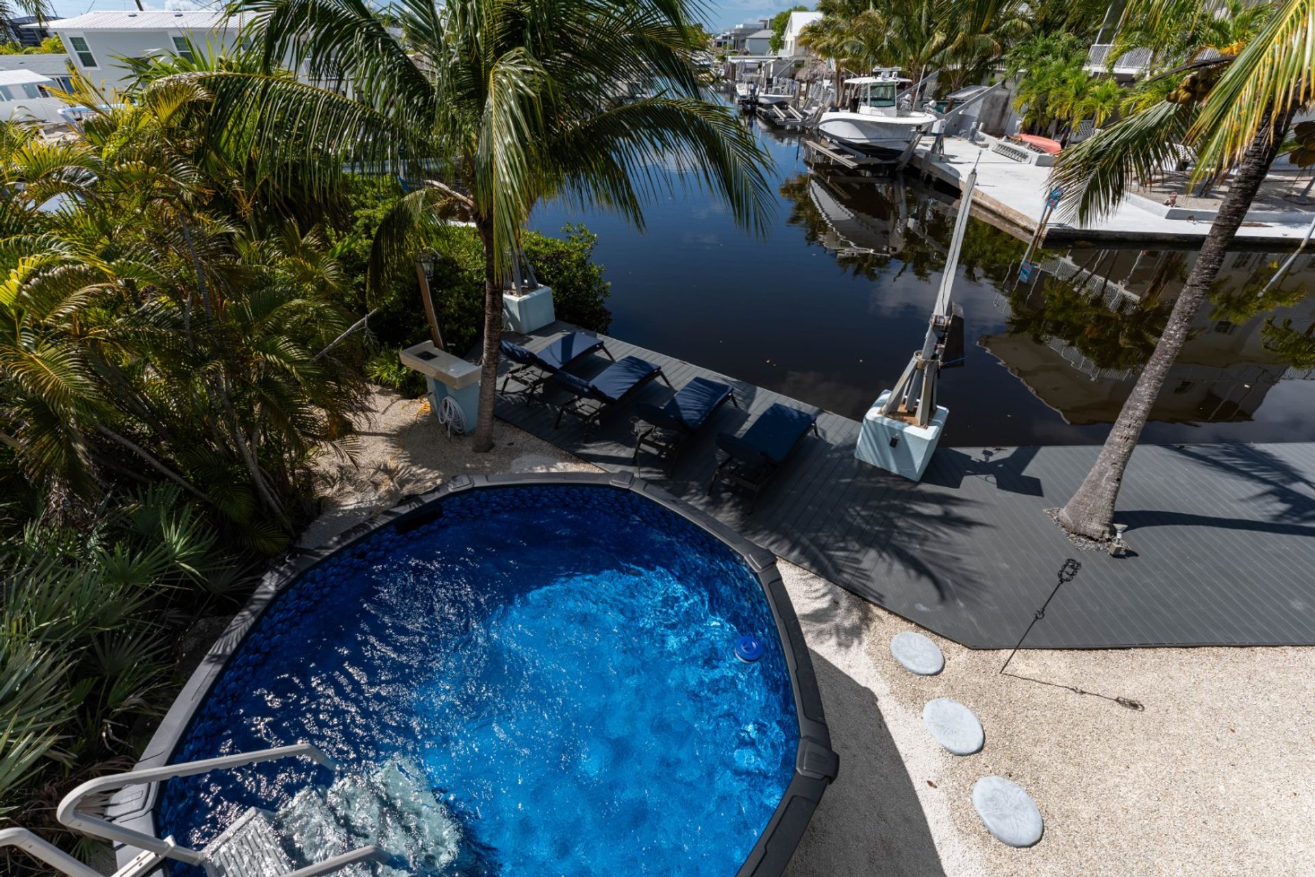 Sunny canalfront tropical backyard with a round blue plunge pool, palm trees, gray dock with lounge chairs, and a boat moored on calm water