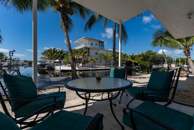 Shaded dockside patio with teal-cushioned chairs around a round glass table, palm trees, boats and a white waterfront house under a bright blue sky.