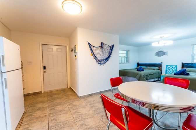 Bright coastal-themed studio interior with tiled floor, retro chrome round table and red diner chairs, two green-made beds with blue accents, fishing-net wall decor and fish art near the entry and fridge