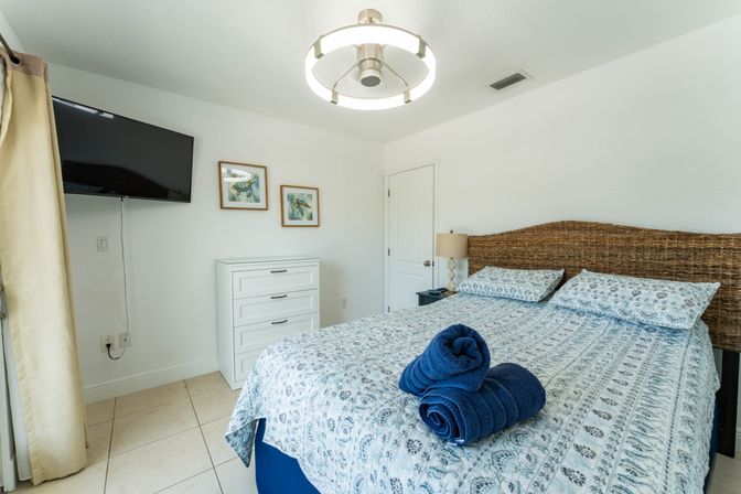 Sunny coastal vacation-rental bedroom with woven rattan headboard, blue patterned bedding, neatly rolled navy towels on the bed, wall-mounted TV, white dresser and sea turtle prints.