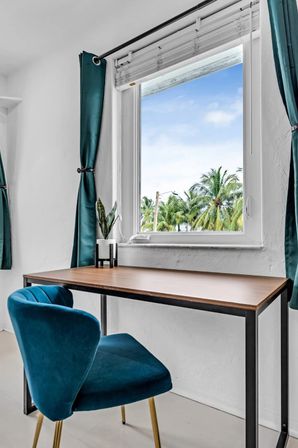Cozy home office desk by a window with teal velvet chair and curtains, small potted plant on the wooden desk and tropical palm-tree view outside.