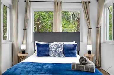 Sunlit modern bedroom with gray headboard, white linens and deep-blue pillows and throw, rolled towels on the bed, wooden nightstands with lamps, beige curtains and large windows overlooking lush green garden.