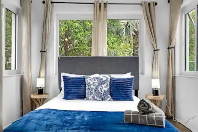 Sunlit modern bedroom with gray headboard, white linens and deep-blue pillows and throw, rolled towels on the bed, wooden nightstands with lamps, beige curtains and large windows overlooking lush green garden.