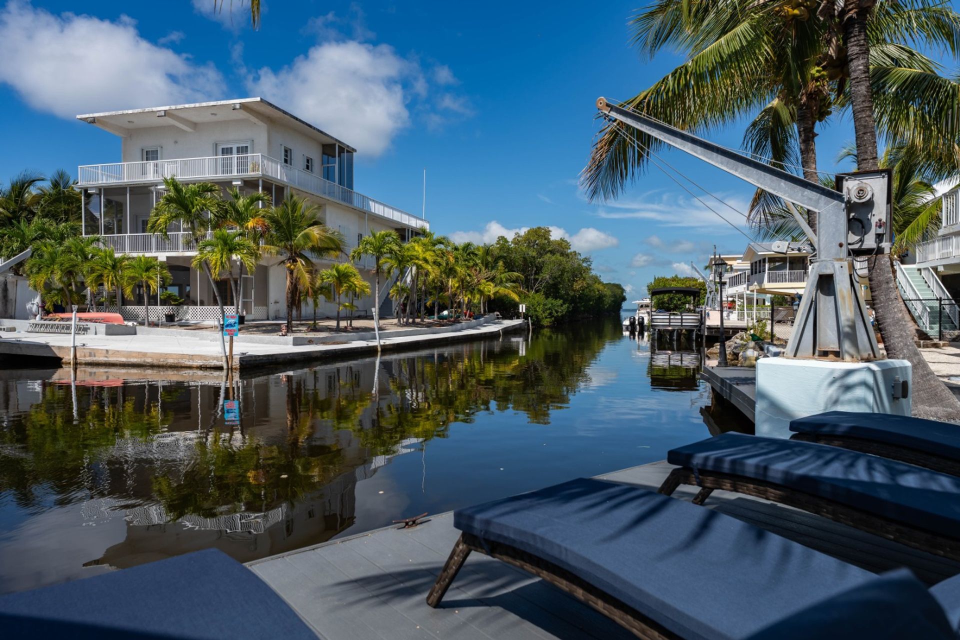Tropical waterfront canal scene with a white multi-story house, palm trees along a concrete dock, boat lift and small boats, and blue-cushioned sun loungers in the foreground under a bright blue sky.