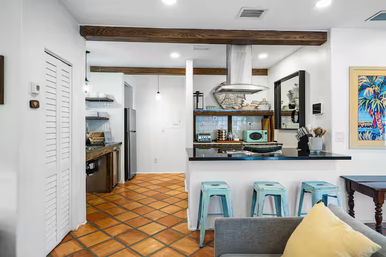 Open-concept kitchen and living area with terracotta tile floor, exposed wooden beams, stainless steel range hood, black countertop with three turquoise metal bar stools, gray sofa with yellow pillow, and colorful tropical wall art.