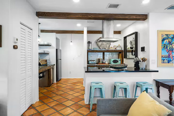 Open-concept kitchen and living area with terracotta tile floor, exposed wooden beams, stainless steel range hood, black countertop with three turquoise metal bar stools, gray sofa with yellow pillow, and colorful tropical wall art.
