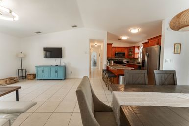 Bright open-plan living and dining area with tiled floors, wall-mounted TV over a turquoise media cabinet, wooden kitchen cabinets, stainless steel refrigerator and a granite island with bar stools.