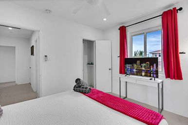 Bright white modern bedroom with a pop of hot-pink curtains and bed runner, rolled towels on the bed, TV on a desk showing a city skyline, and a window view with palm trees.