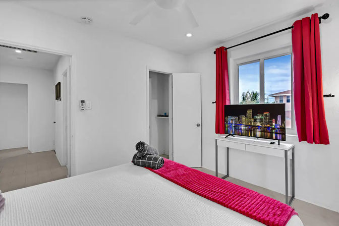 Bright white modern bedroom with a pop of hot-pink curtains and bed runner, rolled towels on the bed, TV on a desk showing a city skyline, and a window view with palm trees.