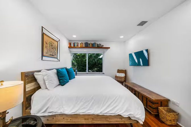 Bright, cozy modern bedroom with white walls and linens, wooden platform bed topped with teal accent pillows, large window looking out to green trees, floating bookshelf, wall-mounted TV, wooden bench and wicker basket.