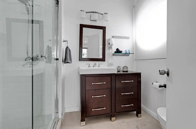 Bright modern bathroom — glass shower, dark wood vanity with white sink and mirror, chrome fixtures, wall shelf with toiletries, towel and sunlit window.