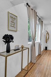 Modern minimalist hallway with white textured walls and hardwood floors, gold-framed glass console table holding a black vase of flowers and small calendar, beige grommet curtains tied at a large window, framed sign reading "OKAY with talking but not touching" and abstract gold floor sculpture.