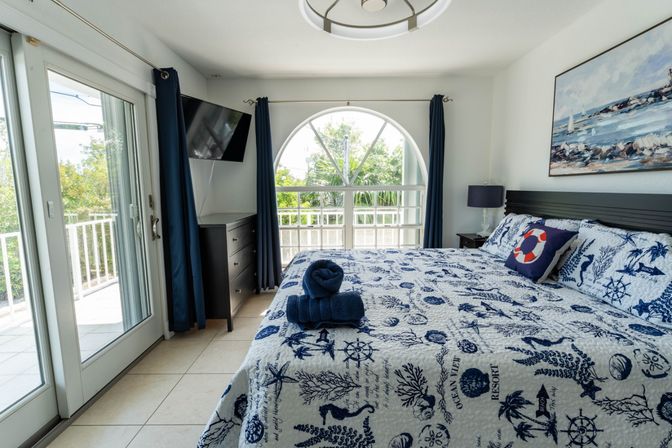 Bright coastal bedroom with king bed in a blue-and-white nautical quilt, life-ring pillow and folded navy towels, sliding glass doors to a balcony, arched window framing tropical greenery, wall-mounted TV and ocean painting