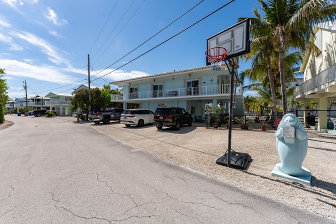 Sunny coastal street with a pastel blue two-story beach house, parked SUVs, portable basketball hoop and a whimsical manatee statue holding a mailbox among palm trees