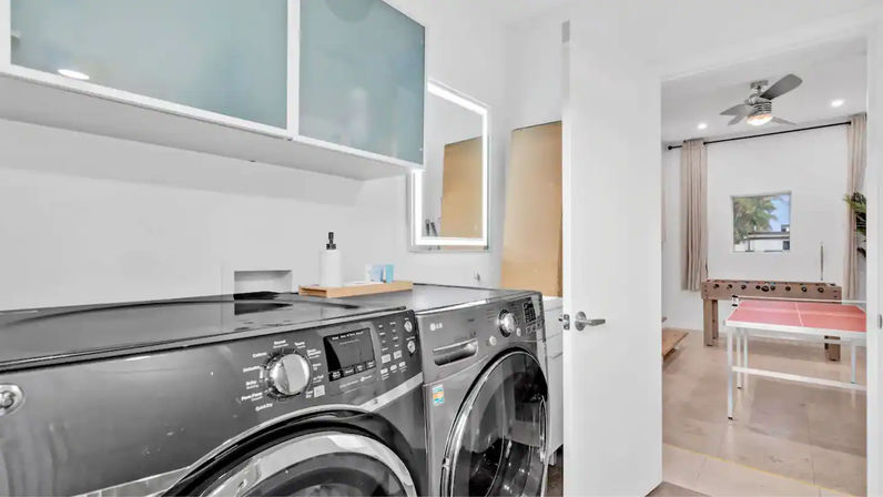 Bright modern laundry room with sleek front-load washer and dryer under frosted cabinets; open door reveals a game room with ping-pong and foosball tables, ceiling fan and a small window with palm trees.