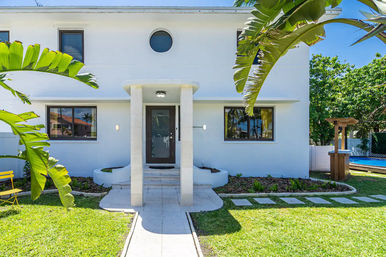 Sunlit modern white two-story house with covered front entry, round window, tropical banana leaves, manicured lawn and backyard pool visible