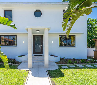 Sunlit modern white two-story house with covered front entry, round window, tropical banana leaves, manicured lawn and backyard pool visible