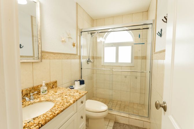 Bright coastal-style small bathroom with beige tile, granite vanity and sink, white toilet, and glass-enclosed walk-in shower with arched window and starfish wall decor