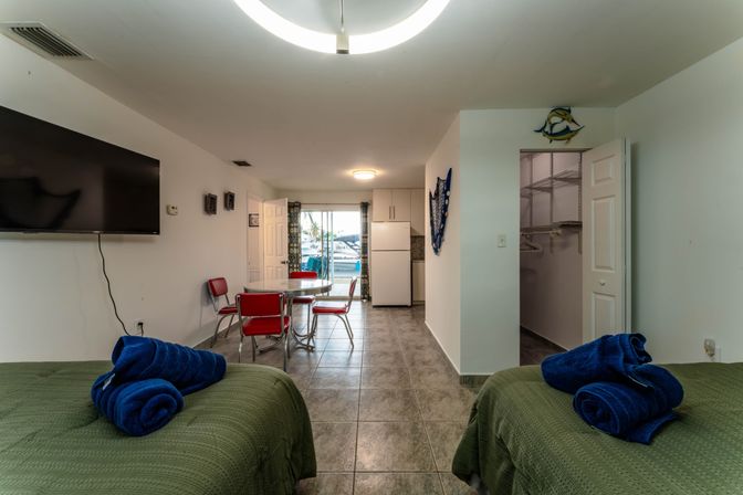 Bright marina-side studio with two twin beds in green covers topped with rolled blue towels, tiled floor, wall-mounted TV, chrome round table with red retro chairs, small kitchenette and open closet, sliding glass door revealing boats at the dock.
