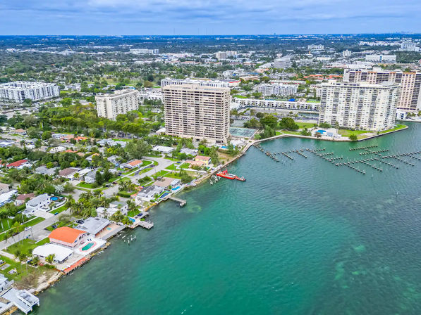 Aerial drone view of a coastal city waterfront with high-rise condo towers, marina pilings, seaside homes and pools along a turquoise bay under a cloudy sky.