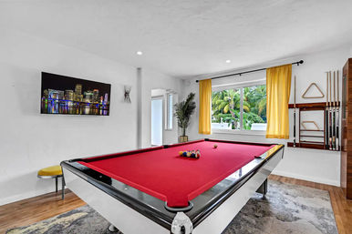 Bright game room with a red-felt pool table, yellow curtains framing a window with palm trees outside, wall-mounted TV showing a city skyline, cue rack, stool and potted plant — modern tropical interior.