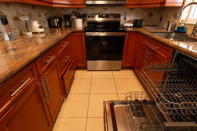 Cozy residential kitchen with open dishwasher rack in foreground, stainless-steel oven, granite countertops, warm wood cabinets, tiled floor and small countertop appliances.