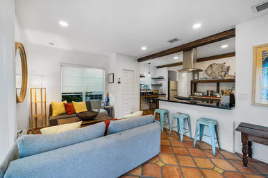 Bright open-plan living room and kitchen with Mediterranean-style terracotta tiles, a light blue sofa with colorful throw pillows, a breakfast bar with three turquoise metal stools, exposed wooden beam and decorative fish wall art — cozy coastal-inspired interior.