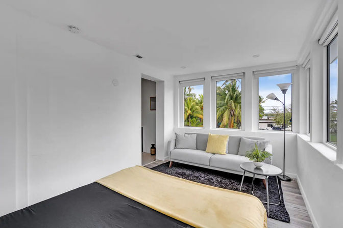 Sunlit modern bedroom with black bedspread and yellow throw, gray sofa, side table and floor lamp beside three windows framing tropical palm trees outside.