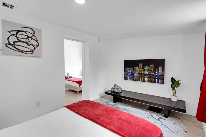 Bright modern urban bedroom with white walls, red bed throw, wall-mounted TV showing a glowing city skyline, low dark bench with rolled towels and potted plant, doorway to a second bedroom.