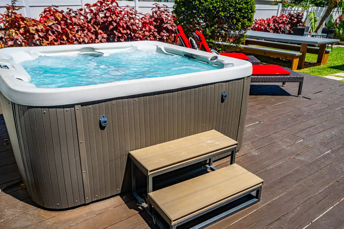Sunny backyard hot tub bubbling on a wood deck with two-step entry, red-cushioned lounge chairs and a picnic table amid tropical plants