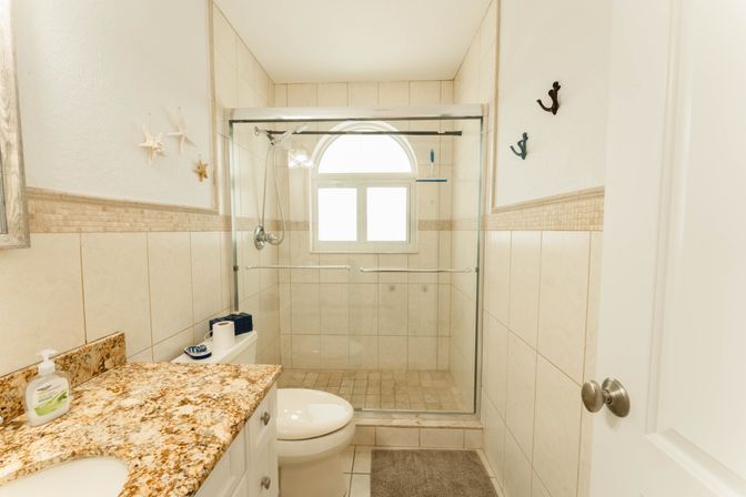 Bright coastal-style bathroom with granite countertop and white vanity, toilet, and glass-enclosed tiled shower with an arched window and starfish wall decor