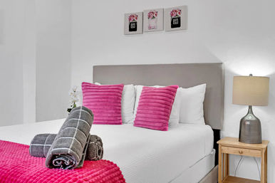 Bright modern bedroom with white walls and gray headboard, two ribbed hot-pink throw pillows on crisp white bedding, rolled gray towels on a pink throw, wooden nightstand with silver lamp and small floral wall art above
