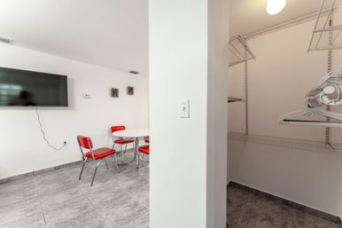 Bright dining nook with retro red chrome chairs around a white round table, wall-mounted flat-screen TV and gray tile floor, next to an open walk-in closet with white wire shelving and hangers.