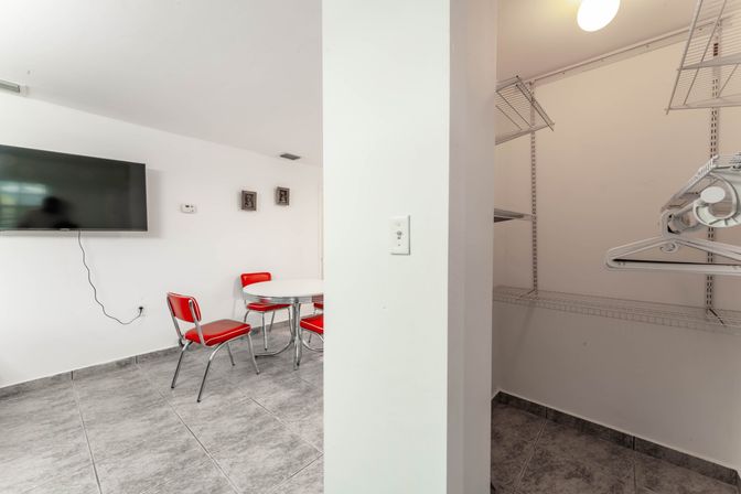 Bright dining nook with retro red chrome chairs around a white round table, wall-mounted flat-screen TV and gray tile floor, next to an open walk-in closet with white wire shelving and hangers.